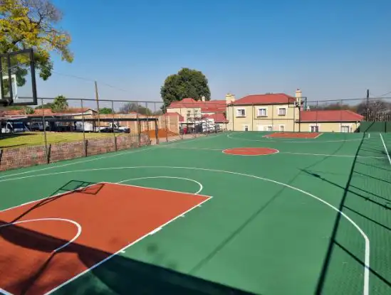 contact-img1 Outdoor basketball court with green and orange surface, white lines, a backboard on the left, and houses in the background on a sunny day.