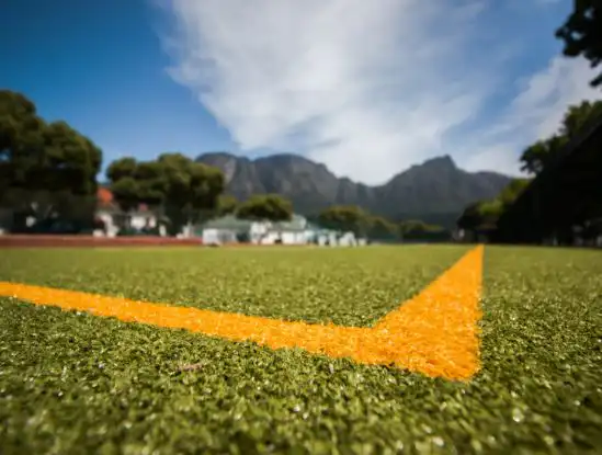 contact-img2 Close-up of bright orange field line forming a corner on artificial turf, with mountains and trees in the background under a blue sky.