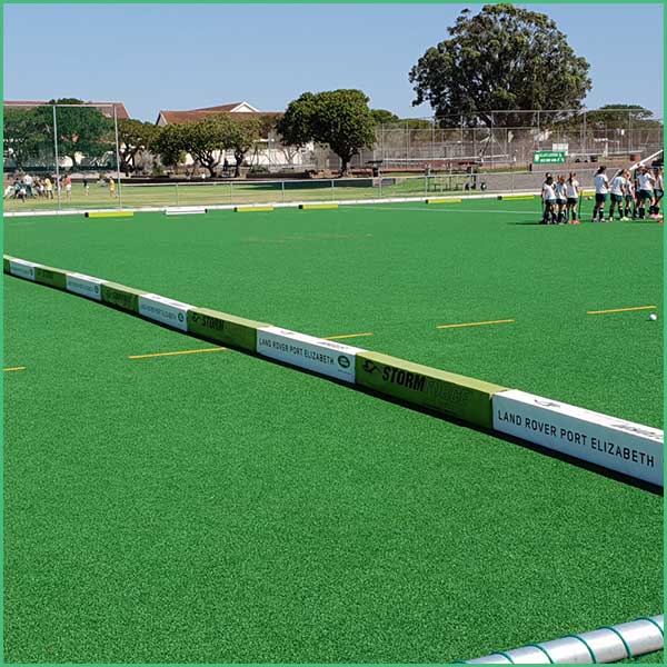 Field hockey practice on bright green artificial turf with a group of players near the sideline in the background