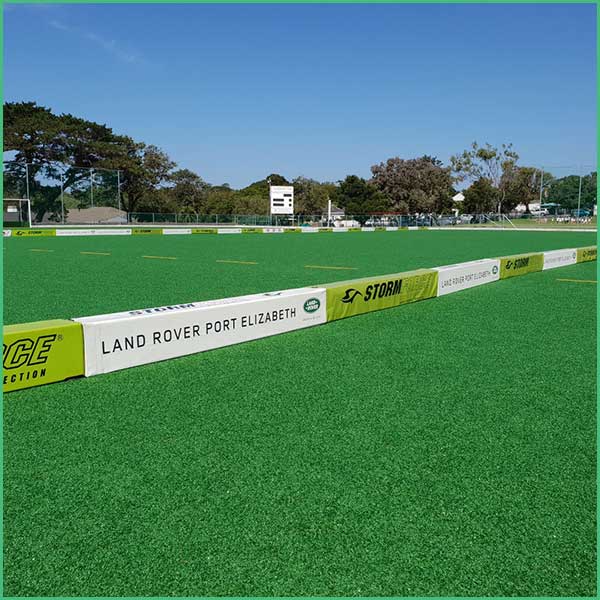 Artificial turf sports field with white and yellow sponsor barriers along the edge and a blue sky behind Trees line the background