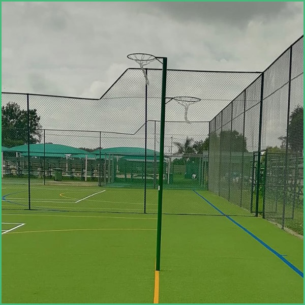 Outdoor fenced sports court with a green surface and blue and yellow boundary lines, enclosed by tall mesh fences and poles on a cloudy day, likely for padel/basketball