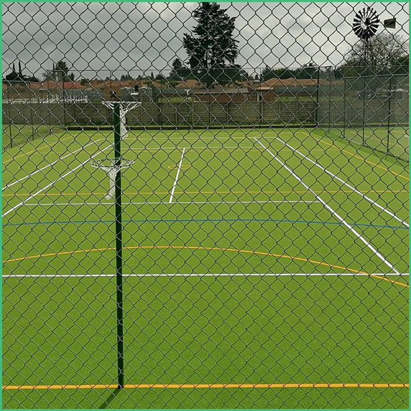Tennis court seen through a chain link fence, with a green surface and white boundary lines; a windmill and trees are visible in the distance