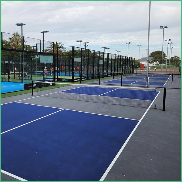 Row of blue outdoor courts with nets and black fencing at a recreation complex, under a cloudy sky