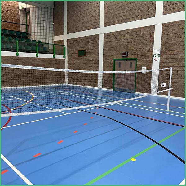 Indoor sports hall with a badminton net across a blue multi use court; green seating and brick walls in the background