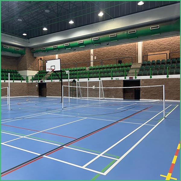 Indoor gym with a badminton net set up on a blue court, green stadium seating along the walls and a basketball hoop in the background