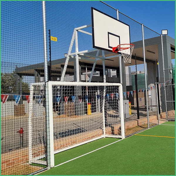 Outdoor sports court with a basketball hoop mounted above a small soccer goal, surrounded by a high chain link fence and festive bunting on a sunny day