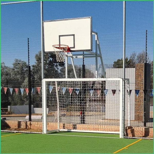 Outdoor sports court featuring a basketball hoop mounted above a small soccer goal on artificial turf, enclosed by a chain link fence with bunting flags