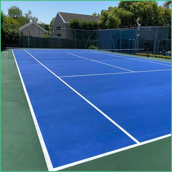 Outdoor blue tennis court with a white net, surrounded by a green fence and residential buildings in the background