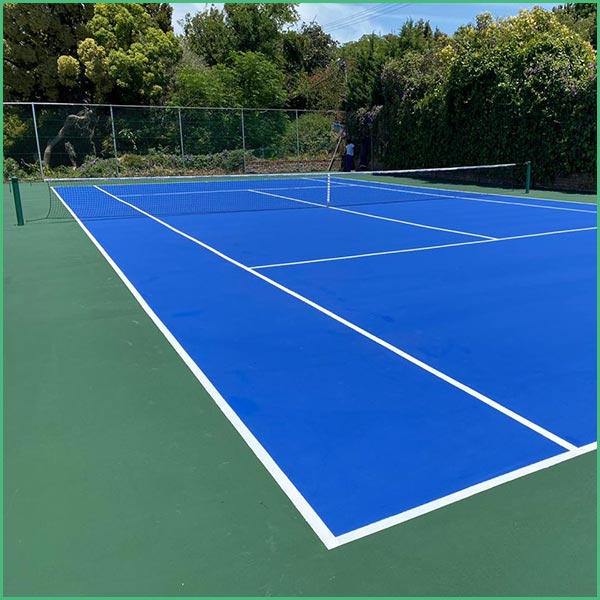 Outdoor blue tennis court with white boundary lines and a net in the distance, surrounded by green trees and bushes