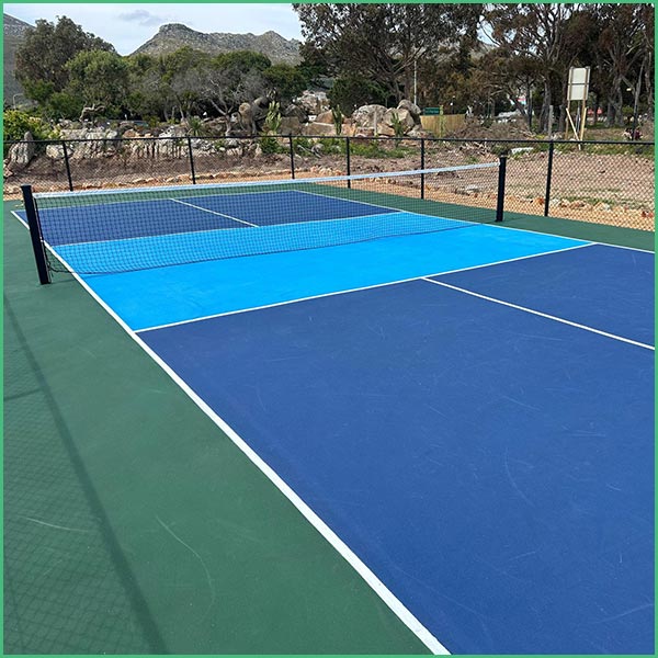 Outdoor blue tennis court with white boundary lines and a net, surrounded by a fence and trees/coastal landscape in the background