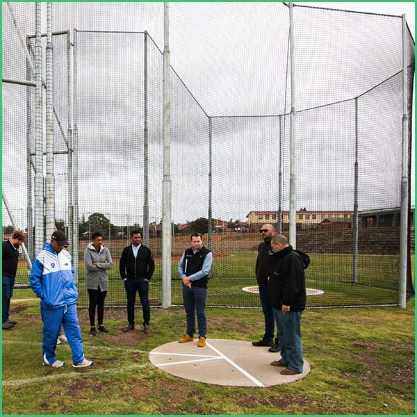 Group of people standing in a fenced baseball/softball practice cage outdoors, around a circular pitch area marked on the ground