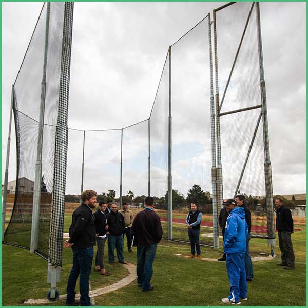 Group of people standing in a circular briefing inside a large outdoor batting cage with tall netting and metal frames on a sports field in cloudy weather 