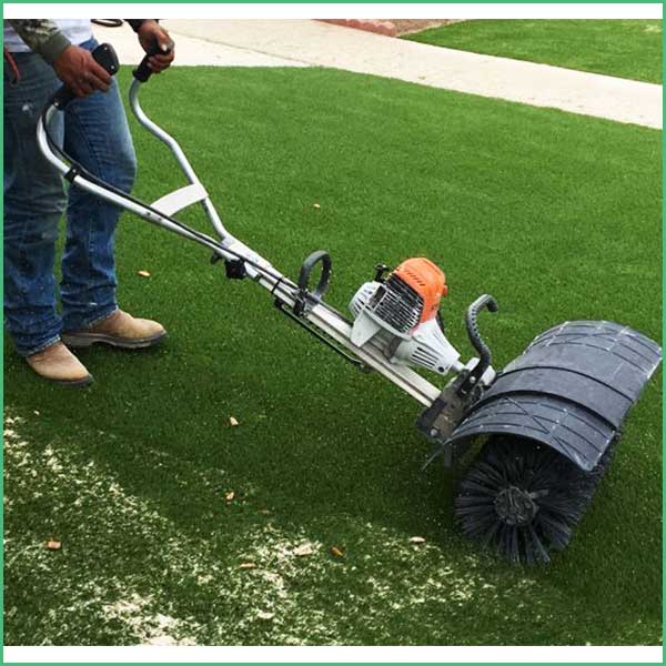 Person operating a gas powered lawn aerator with a large cylindrical roller across a green lawn near a sidewalk