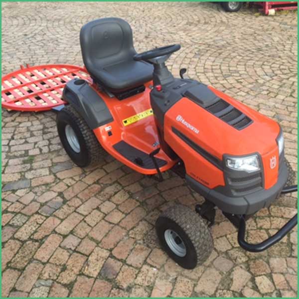 Orange riding lawn mower with black seat on a cobblestone driveway, front headlights visible and a red rear attachment attached behind it