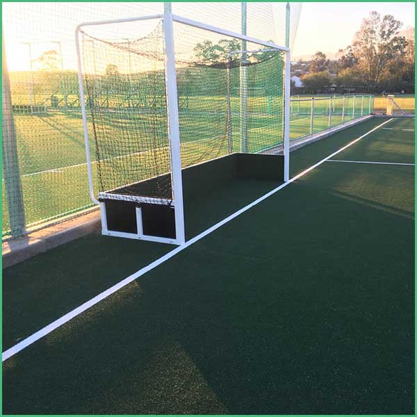 Small soccer goal with a built in bench at the front on a green turf field, surrounded by a high fence and white boundary lines at sunset