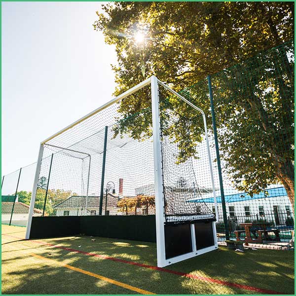 Outdoor sports court with a white metal goal and netting, sun filtering through trees above a fenced turf field