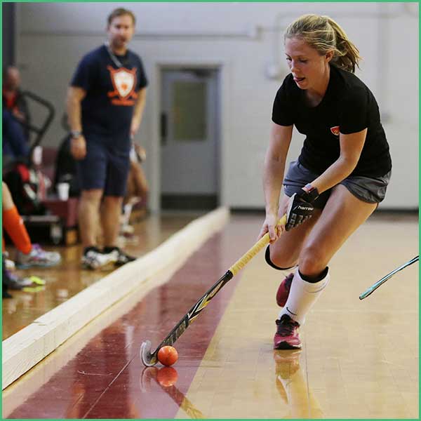 Female floor hockey player in a black shirt and gray shorts maneuvers an orange ball with a stick on a gym floor, with a man watching in the background