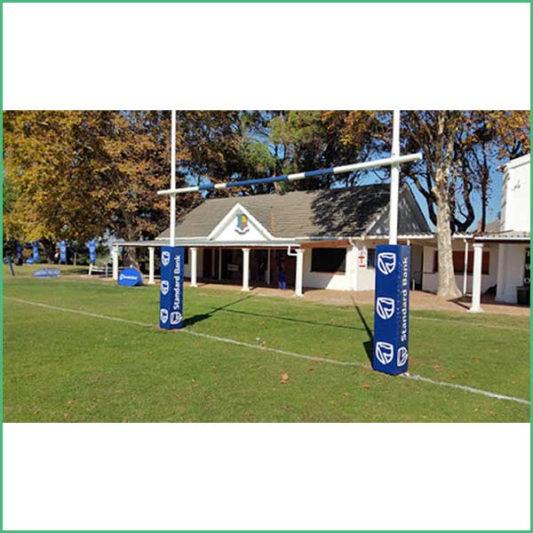 Rugby goal posts on a grassy field with blue padded supports and a clubhouse in the background, autumn trees nearby