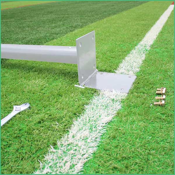 Metal base plate and vertical post anchored to the ground on a grassy sports field, with a white sideline nearby; a wrench and bolts sit on the grass next to the setup