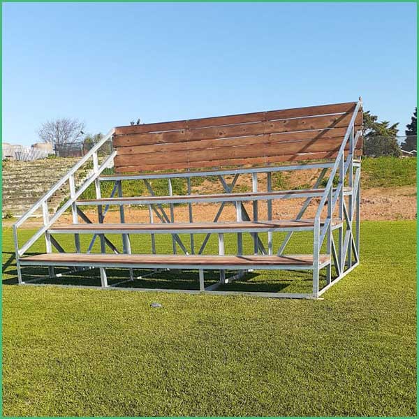 Outdoor metal bleachers with wooden seats on a grassy field, with stairs on the left and a clear blue sky behind