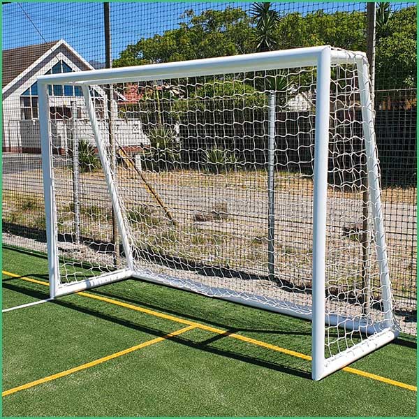White metal soccer goal with net on a green artificial turf; yellow court lines and a fence in the background 