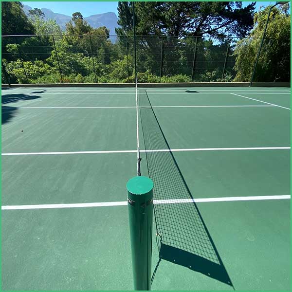 Outdoor tennis court with a net, green surface and white lines, surrounded by trees and distant hills