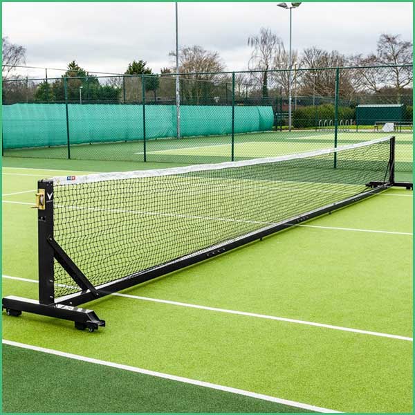 Outdoor tennis court with a net in the foreground and a green windscreen along the fence, sunny day