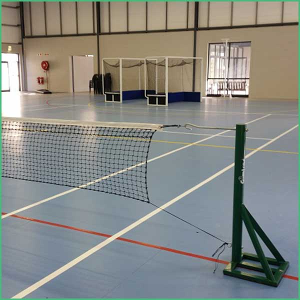 Indoor gym featuring a badminton net across a blue court, with white boundary lines and equipment stored along the back wall