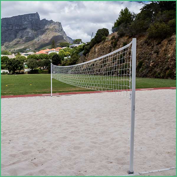 Outdoor beach volleyball court with a white net on sand and a green field beyond, set against rocky hills and distant houses under a cloudy sky