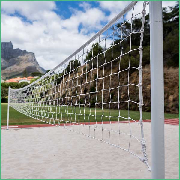 White volleyball net stretched across a sandy court with green grass and hills in the background under a blue sky