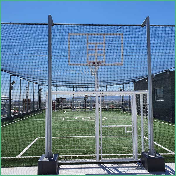 Rooftop fenced basketball court with a hoop and backboard, green artificial turf under a clear blue sky