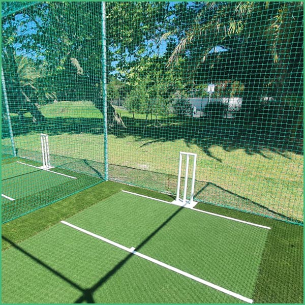 Cricket practice net: green turf lanes with white crease lines and two wicket stumps, enclosed by green netting in a sunny outdoor field