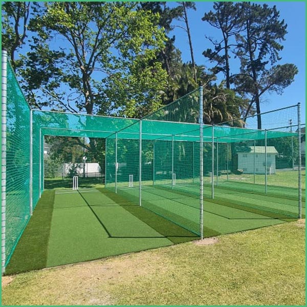 Cricket practice nets with green mesh fencing and artificial turf inside a fenced cage, trees in the background