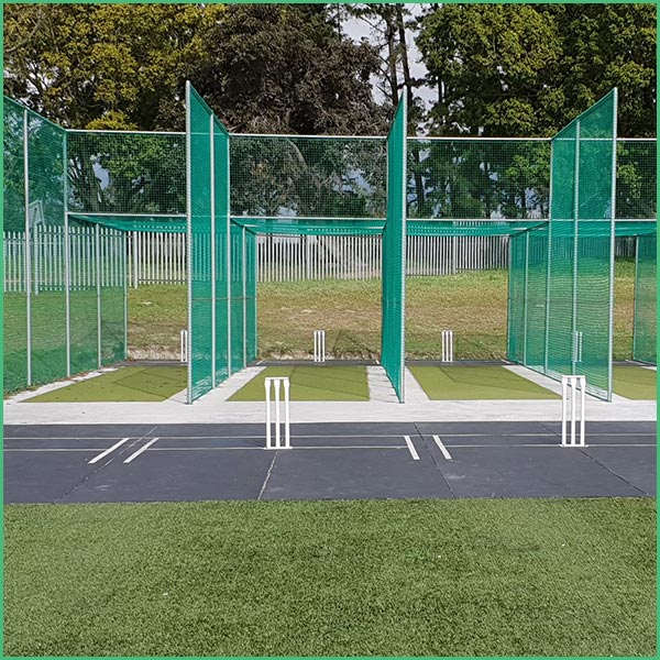 Cricket practice nets with tall green mesh screens and three batting bays, white stumps on artificial pitches, outdoors on a sunny day